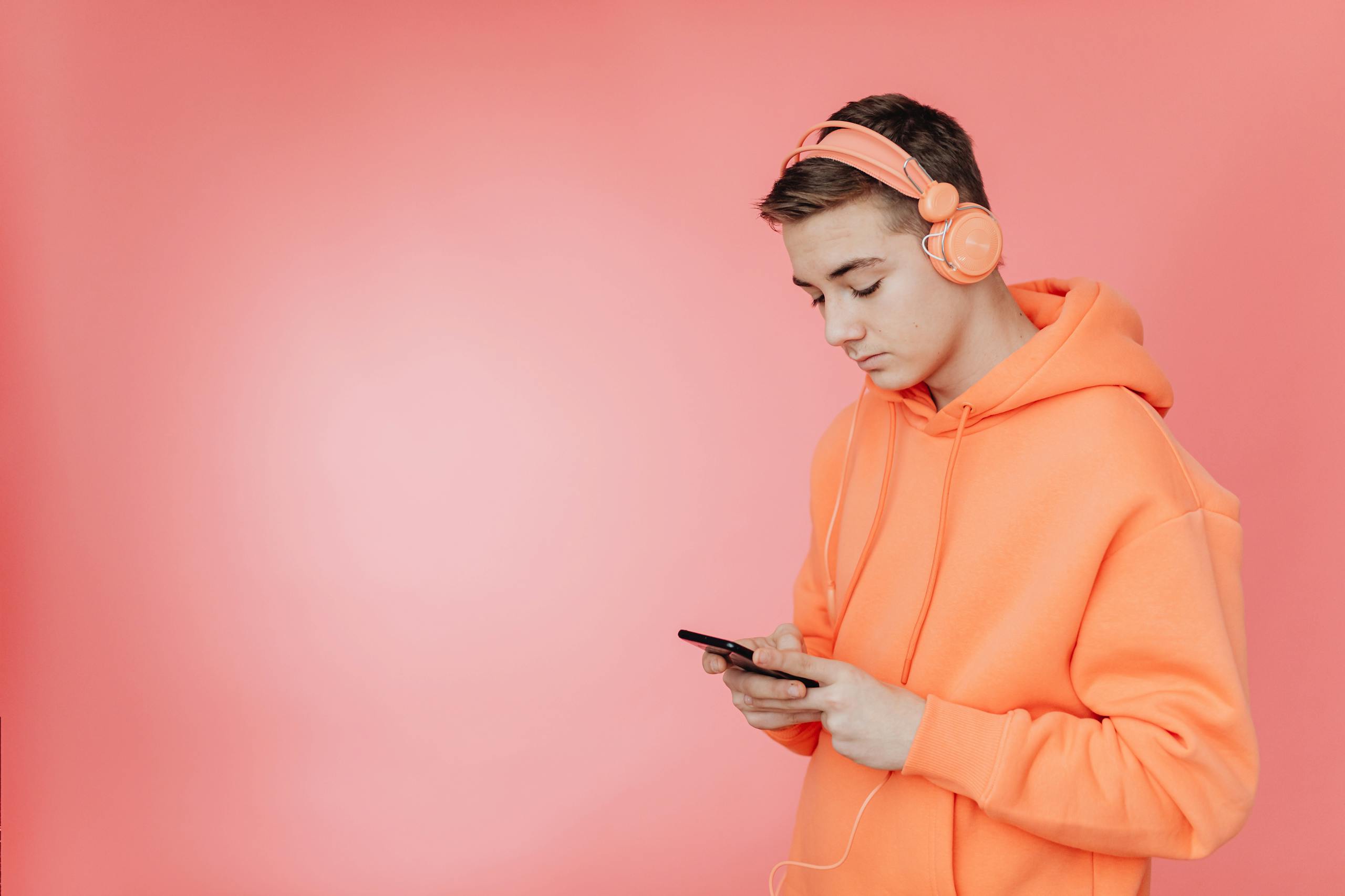 Teenager in orange hoodie using smartphone and headphones against pink backdrop